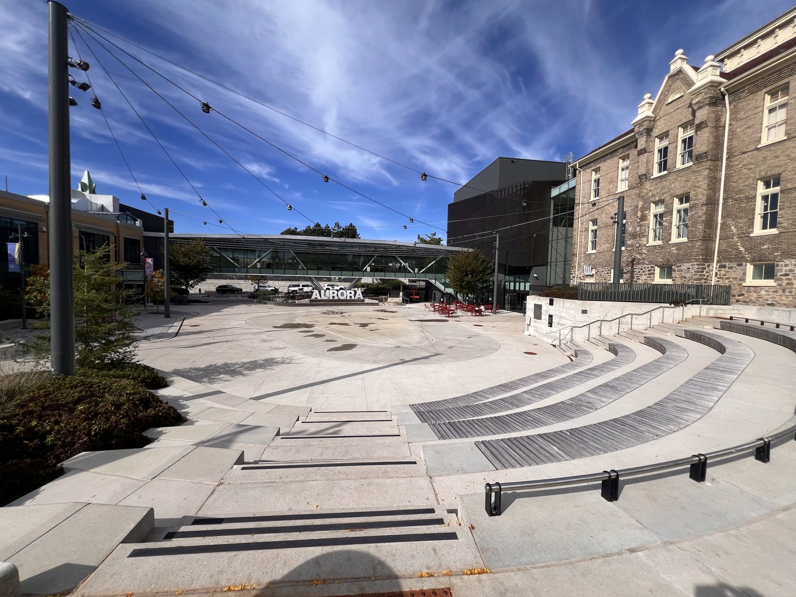 Aurora Town Square: curved amphitheatre steps facing an open plaza, the white AURORA sign and the historic Church Street School under a blue autumn sky